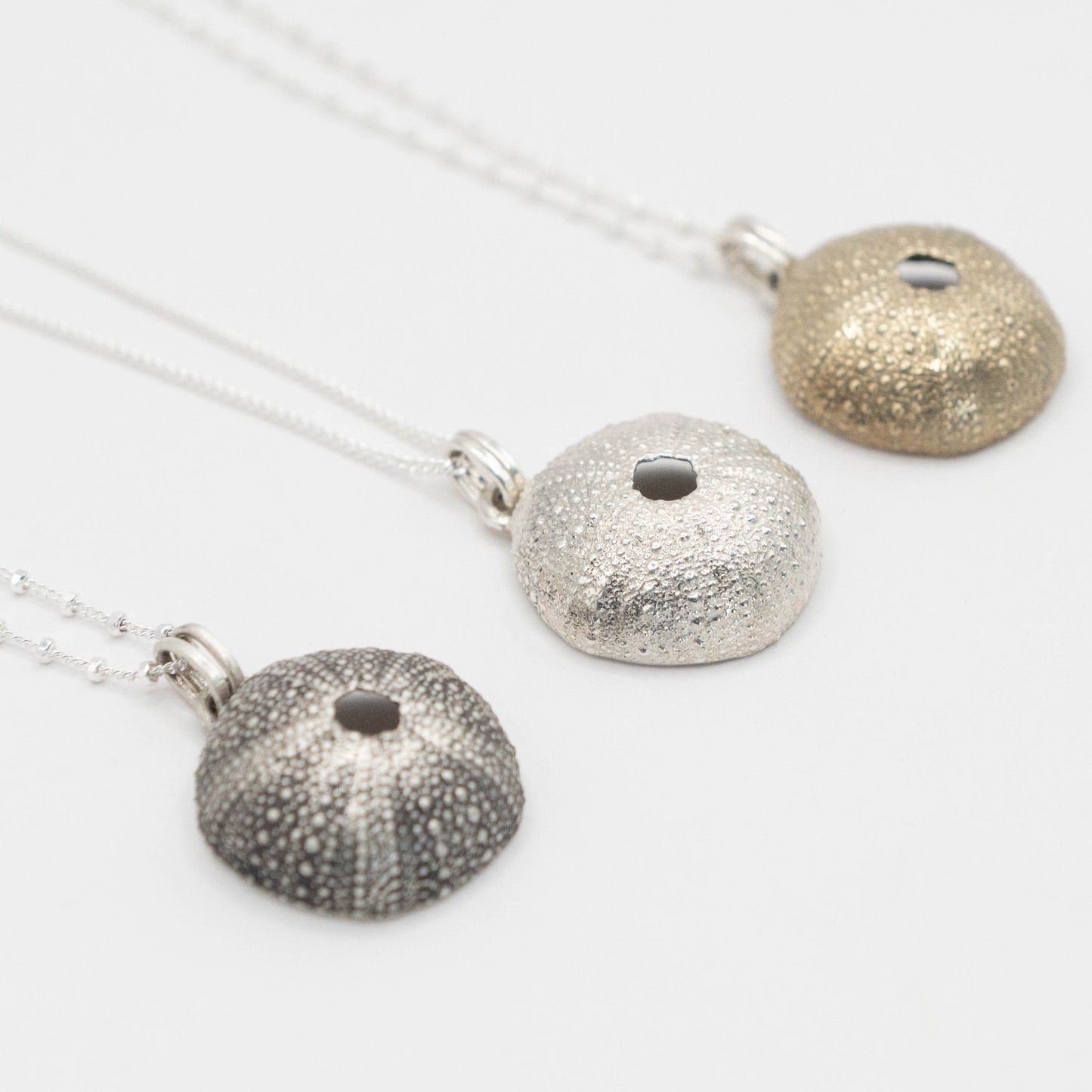 Three sea urchin pendants in oxidized silver, shiny silver, and brass, displayed side by side. Each pendant features the intricate textured surface of a sea urchin shell, hanging from fine silver chains