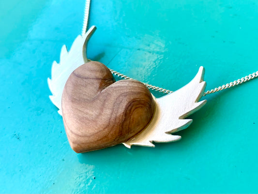 Olive wood heart pendant with sterling silver wings on a turquoise background, reflecting bespoke craftsmanship and a love for natural materials.