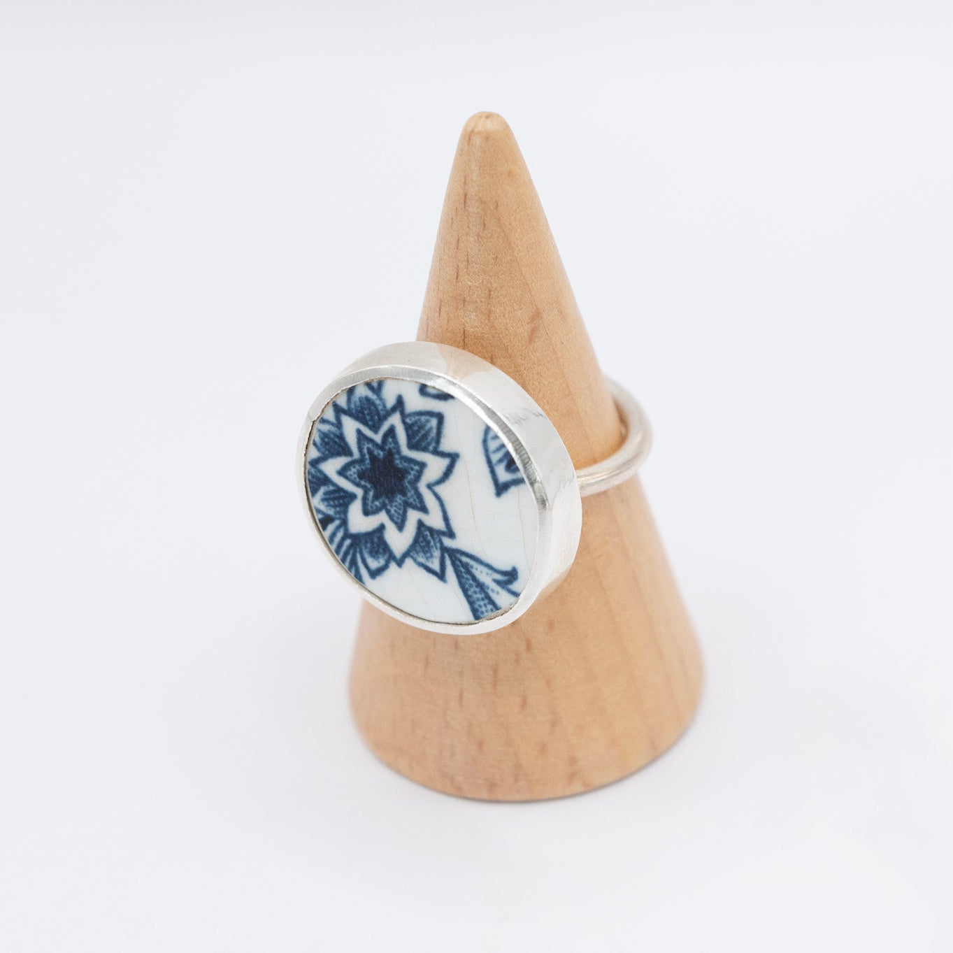 A blue and white antique ceramic ring with a floral design, set in a sterling silver band, displayed on a wooden stand.