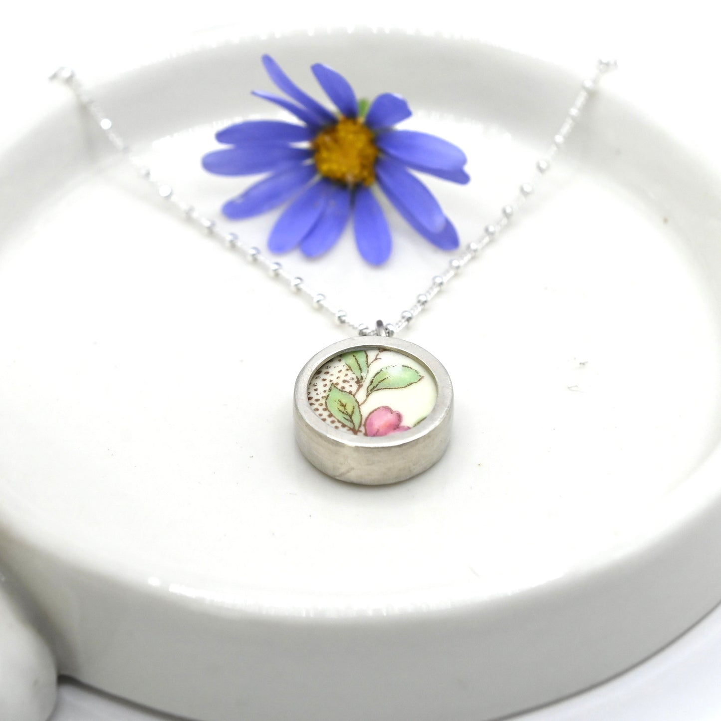 ceramic and silver pendant, presented on a white dish, showing the pink and green floral design on the ceramic and side of the silver bezel setting with a chain.