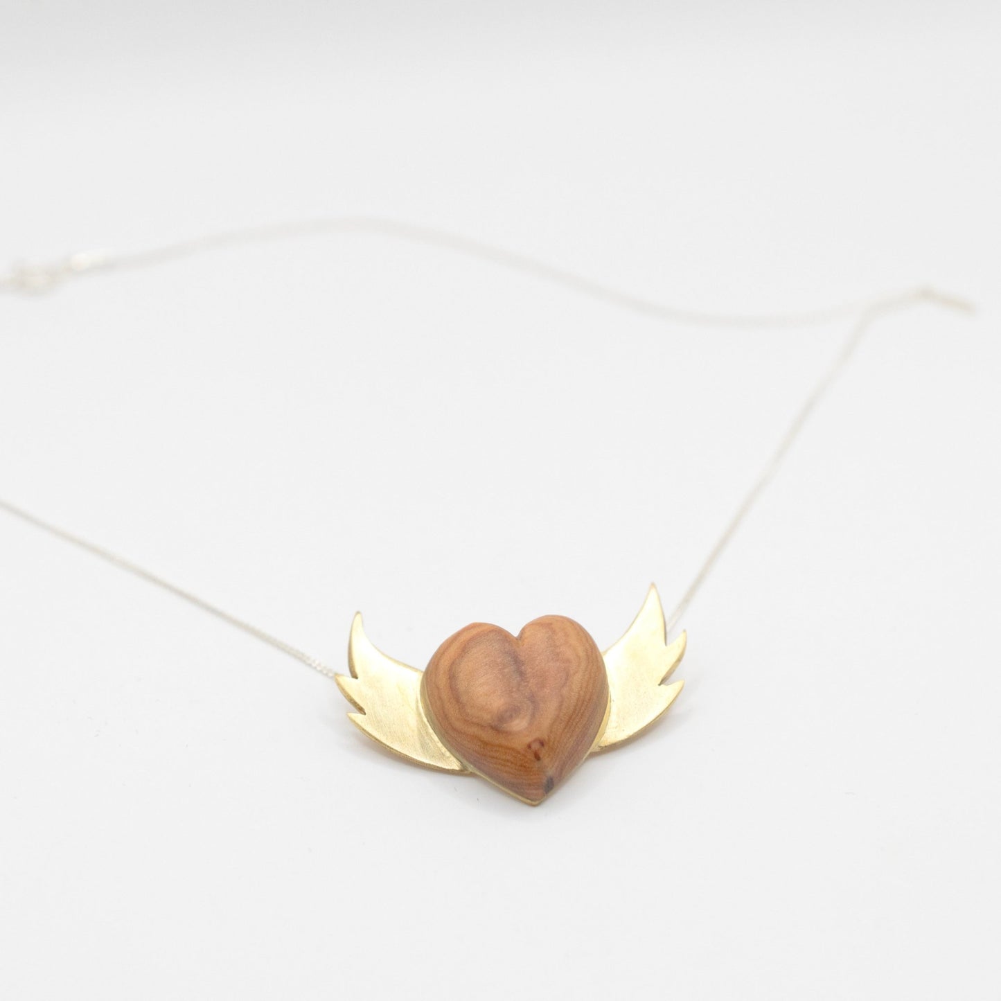 A necklace featuring a heart-shaped pendant made of wood, with a small blue dot at the center. The heart is flanked by brass wings on either side, all attached to a thin silver chain.