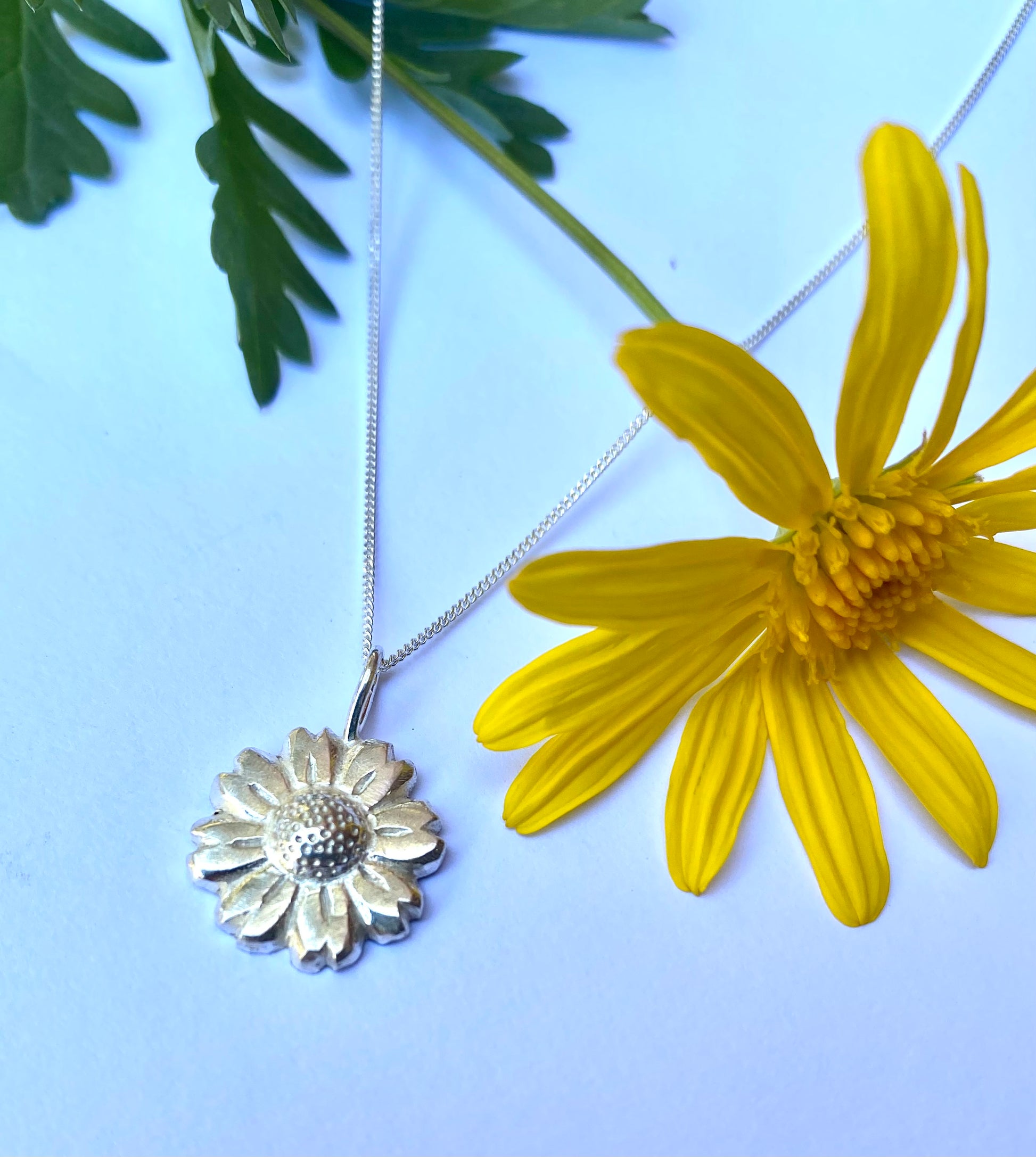 Close-up of shiny Daisy Flower pendant against a fresh yellow daisy backdrop, highlighting the pendant’s detail.