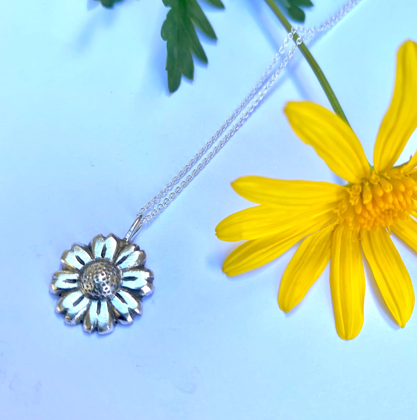Sterling silver Daisy Flower pendant showcased alongside a real yellow daisy on a light blue background