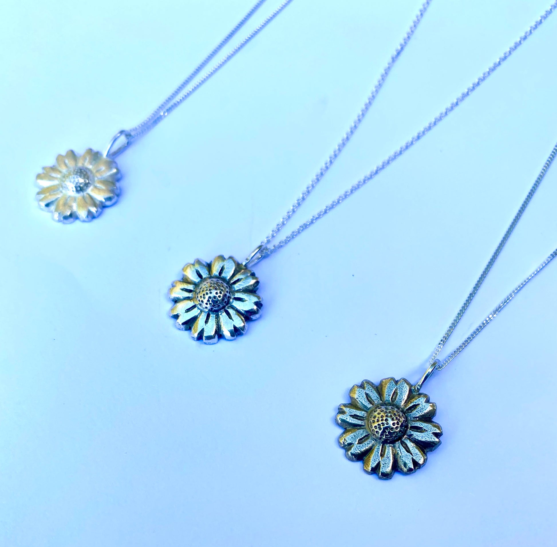 Trio of Daisy Flower pendants in shiny, oxidized, and brass finishes displayed on light blue surface.