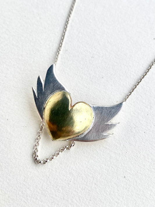 Handcrafted pendant with a glossy brass heart and textured silver wings against a clean white background, showcasing the pendant's fine details and mixed metal design