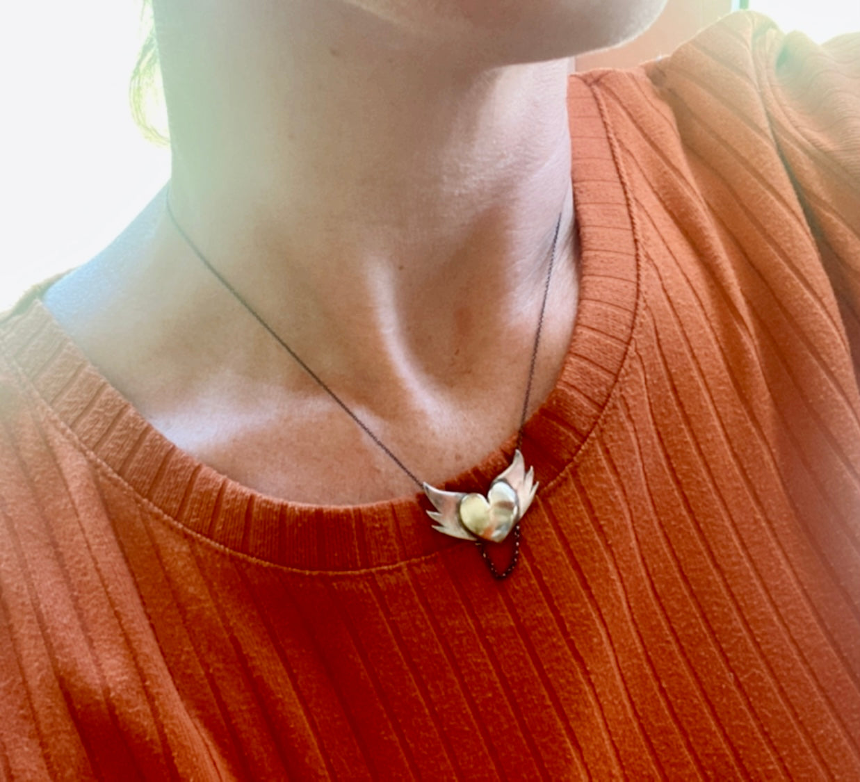 The silver winged brass heart pendant worn on a model, showing how it elegantly drapes around the neck with a partial view of an orange blouse.