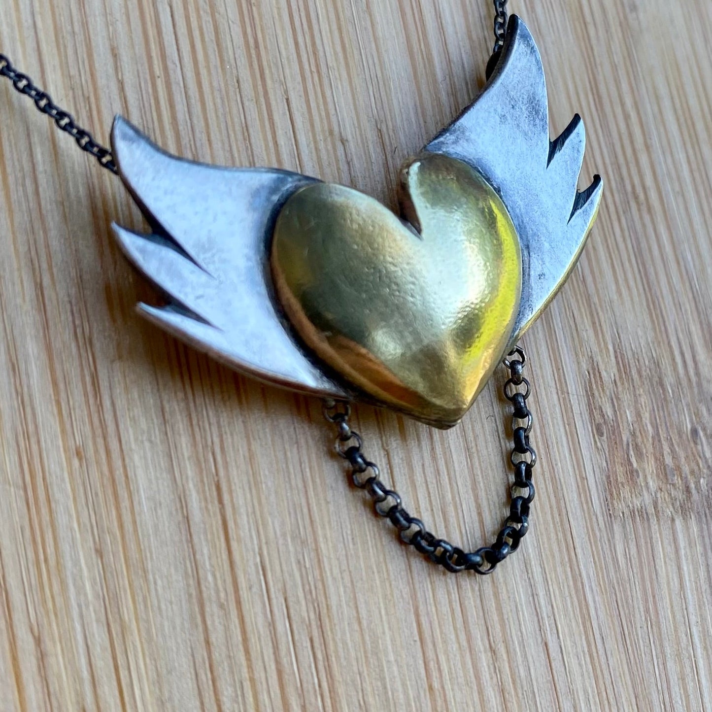 Close-up of the silver winged brass heart pendant laid flat, highlighting the contrast between the shiny brass and the matte silver finishes.