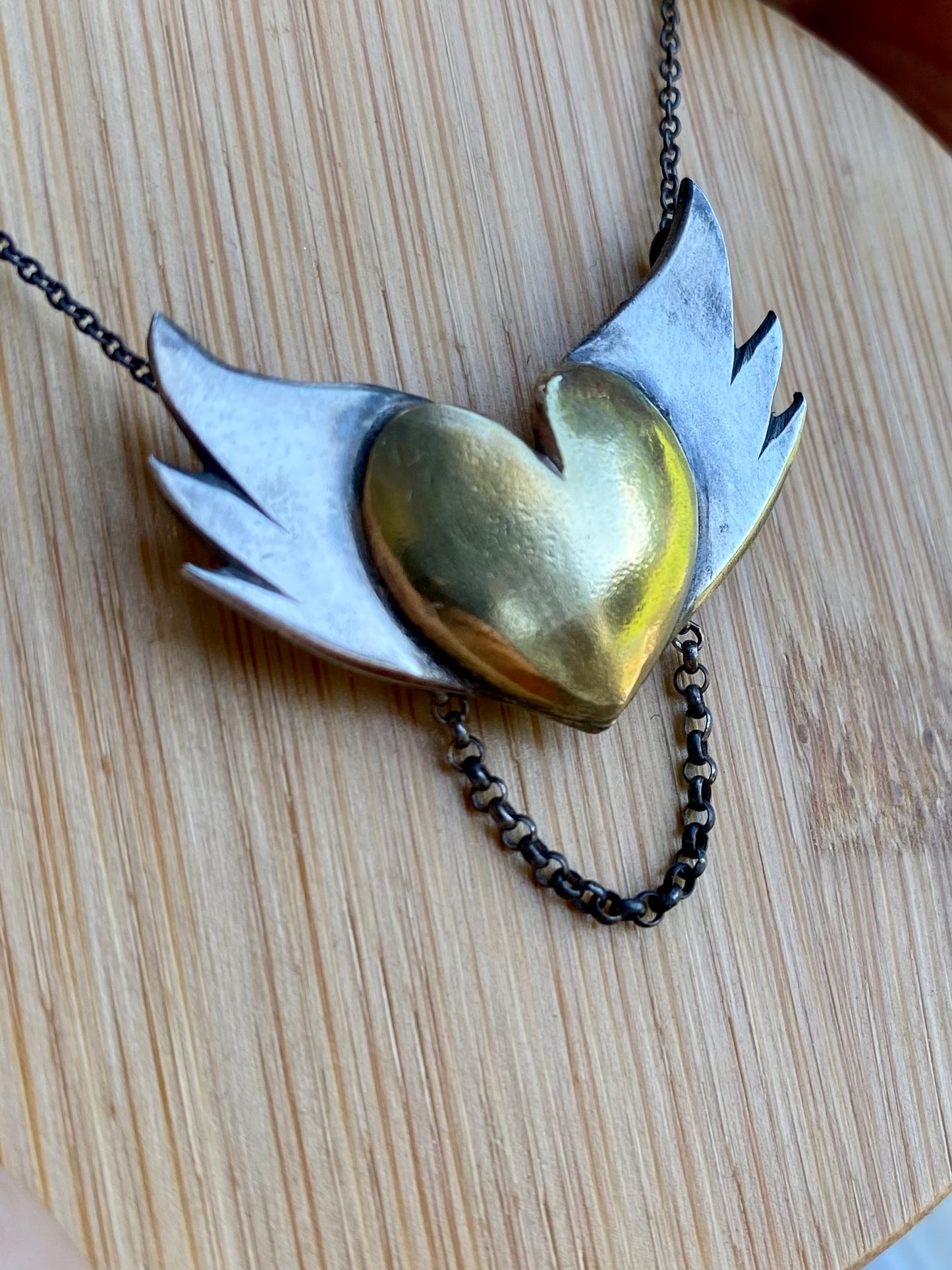 Close-up of the silver winged brass heart pendant laid flat, highlighting the contrast between the shiny brass and the matte silver finishes.