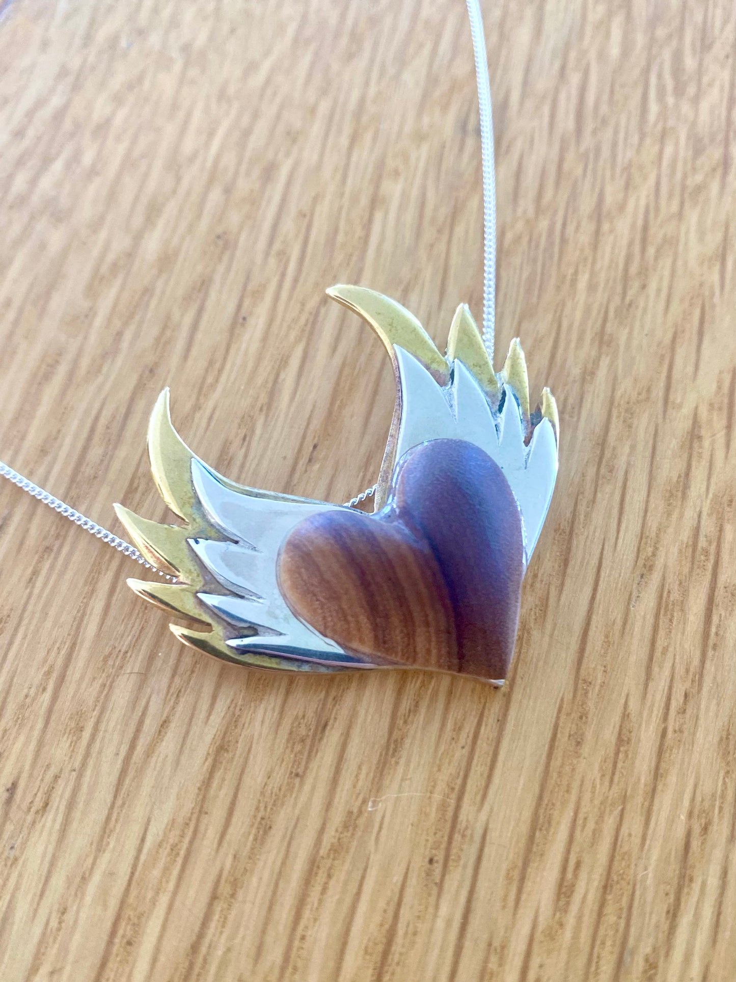 Handcrafted pendant featuring an olive wood heart with silver wings on a light wooden background.