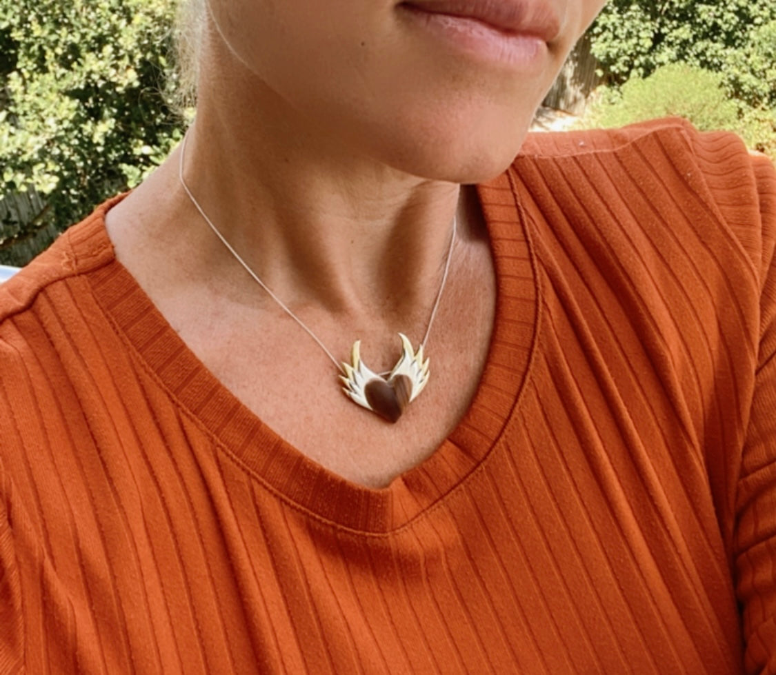 The olive wood heart pendant with silver wings worn by a person in an orange top, showcasing how it looks when worn.