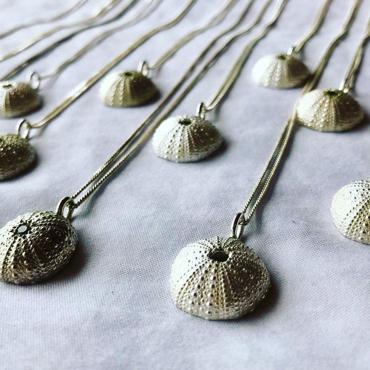 Assortment of sterling silver sea urchin pendants with intricate details on silver chains, laid out on a soft gray background