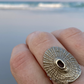 Close-up of a handcrafted brass Santa Maria shell ring worn on a finger against a blurred beach backdrop.
