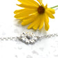 Silver bracelet with a daisy charm next to a yellow flower on a white background