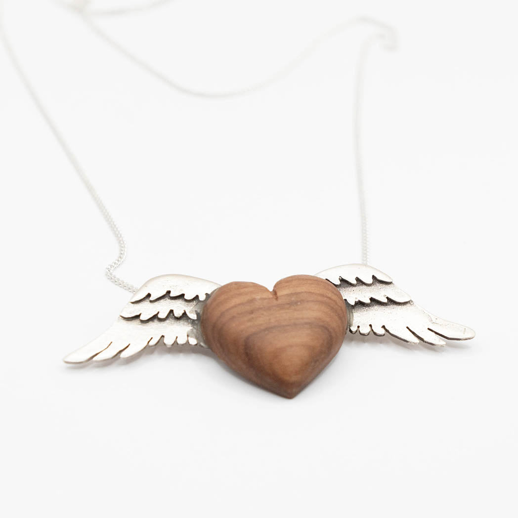 A necklace with a heart-shaped pendant made from olive wood, flanked by silver wings on both sides. The pendant hangs from a delicate silver chain, combining natural wood texture with sleek metallic elements