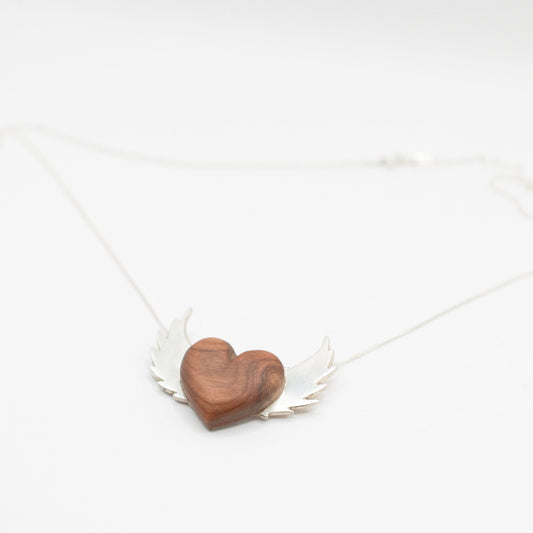 A necklace featuring a heart-shaped pendant made of wood. The heart is flanked by silver wings on either side, all attached to a thin silver chain.