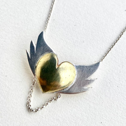 Handcrafted pendant with a glossy brass heart and textured silver wings against a clean white background, showcasing the pendant's fine details and mixed metal design